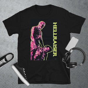 May include: Black t-shirt featuring a graphic illustration with a skeleton figure and the text "HELLRAISER" in yellow. The artwork depicts a skeletal figure and another person in a violent pose. A pair of black sneakers, a phone, and headphones are also visible.