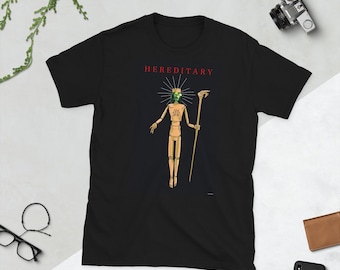 May include: Black t-shirt featuring the word "HEREDITARY" in red above a graphic of a wooden figure with a green face, crown, and staff. The figure is in a pose with arms outstretched.