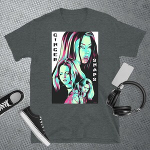 May include: A dark heather grey t-shirt featuring a graphic print of three women in a neon green and pink colour scheme. The text "GINGER SNAPS" is printed vertically on the right side of the graphic.
