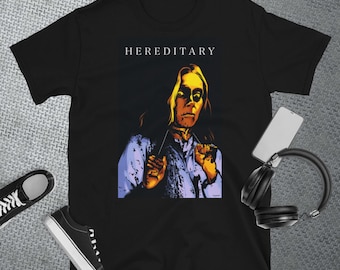 May include: Black t-shirt with a graphic of a person with yellow eyes and a purple shirt. The text "HEREDITARY" is printed in white above the graphic.