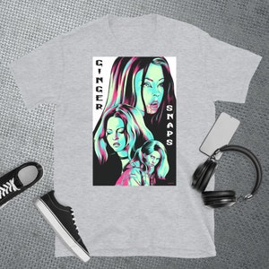 May include: A grey t-shirt featuring a graphic print of three women in a neon green and pink colour scheme. The text "GINGER SNAPS" is printed vertically on the right side of the graphic.