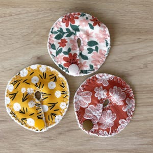 May include: Three round, floral-patterned nursing pads in yellow, red, and white. Each pad has a slit and a button closure. The pads feature various floral designs in pink, white, and green. The pads are made of fabric and are designed for nursing mothers.