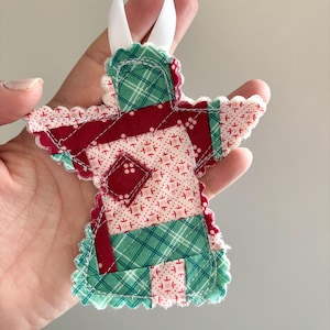 May include: A fabric angel ornament featuring a patchwork design of red, green, and cream-colored cotton fabrics with various patterns. The ornament has scalloped edges with visible white stitching and a white ribbon loop attached to the top.