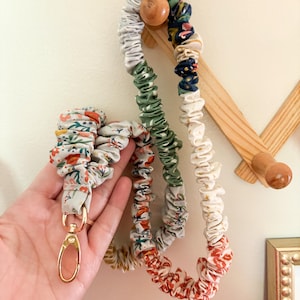 May include: A fabric lanyard featuring a gathered, scrunchie-style design with a multi-colored floral pattern in shades of red, green, blue, and white. The lanyard includes a gold-toned metal swivel lobster clasp at the end for attaching items.