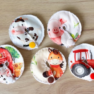 May include: Set of five round, white fabric pads with farm animal and tractor designs. Each pad features a different illustration: a cow, pig, horse, tractor, and a cow with a barn. The pads have a button closure.