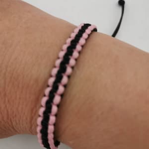 May include: A pink and black macrame bracelet is shown on a wrist. The bracelet is made of woven cord, with alternating rows of pink and black. The adjustable closure is black.