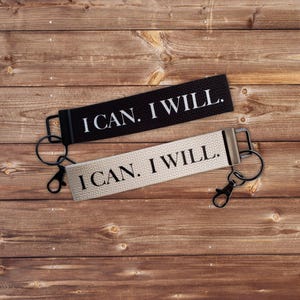 Puede incluir: Dos llaveros con el texto «I CAN. I WILL.» se muestran sobre una superficie de madera. Un llavero es negro con texto blanco, y el otro es blanquecino con texto negro. Cada llavero tiene un anillo y un cierre metálico.