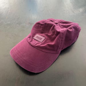 Könnte beinhalten: Eine burgunderfarbene Baseballkappe mit einem kleinen rosa Patch, auf dem vorne "PINK" steht.