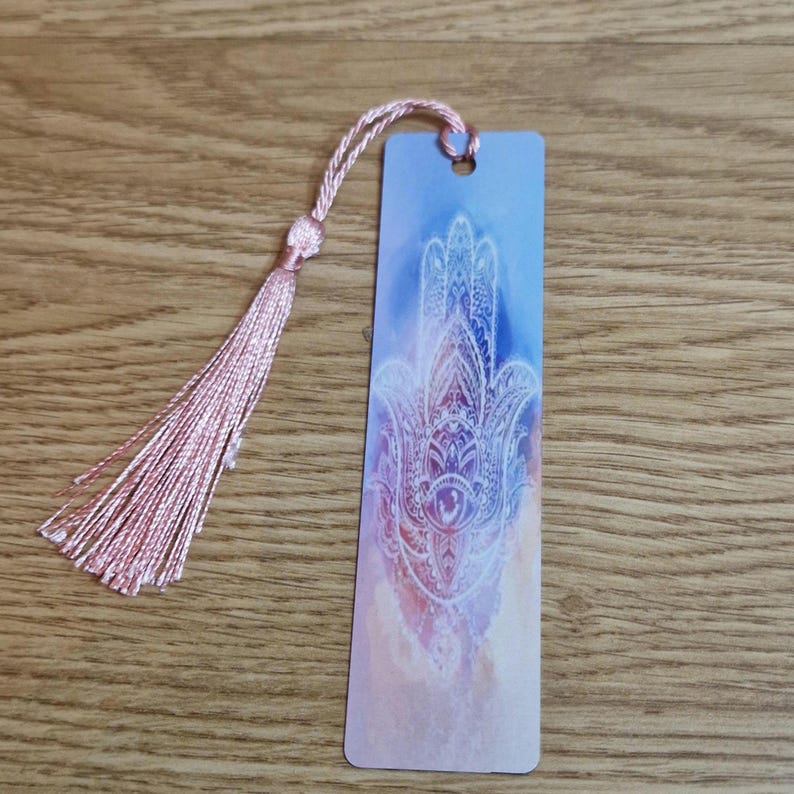 K&ouml;nnte beinhalten: Rechteckiger Lesezeichen mit einem rosa Quastenanh&auml;nger. Das Lesezeichen hat einen blau-rosa Aquarellhintergrund mit einem wei&szlig;en Hamsa-Hand-Design. Die Quaste besteht aus rosa Faden und ist oben am Lesezeichen befestigt.