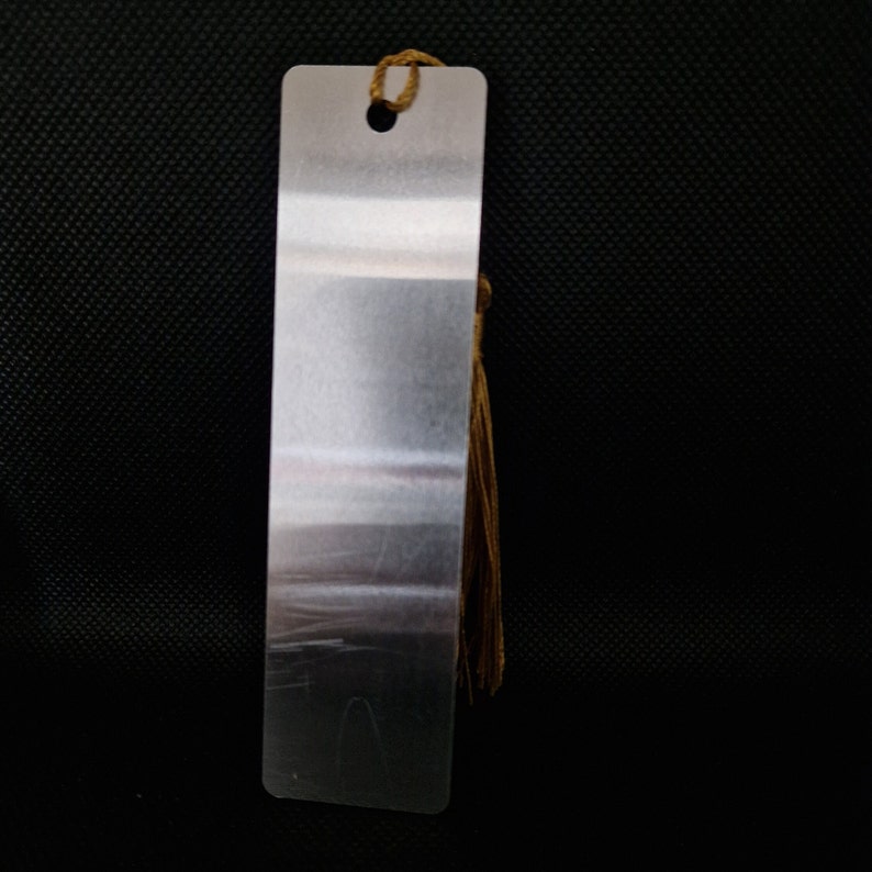 K&ouml;nnte beinhalten: Ein rechteckiger, silberfarbener Metall-Lesezeichen mit einem kleinen Loch oben und einer braunen Quaste. Das Lesezeichen hat eine geb&uuml;rstete Metalloberfl&auml;che vor dunklem Hintergrund.