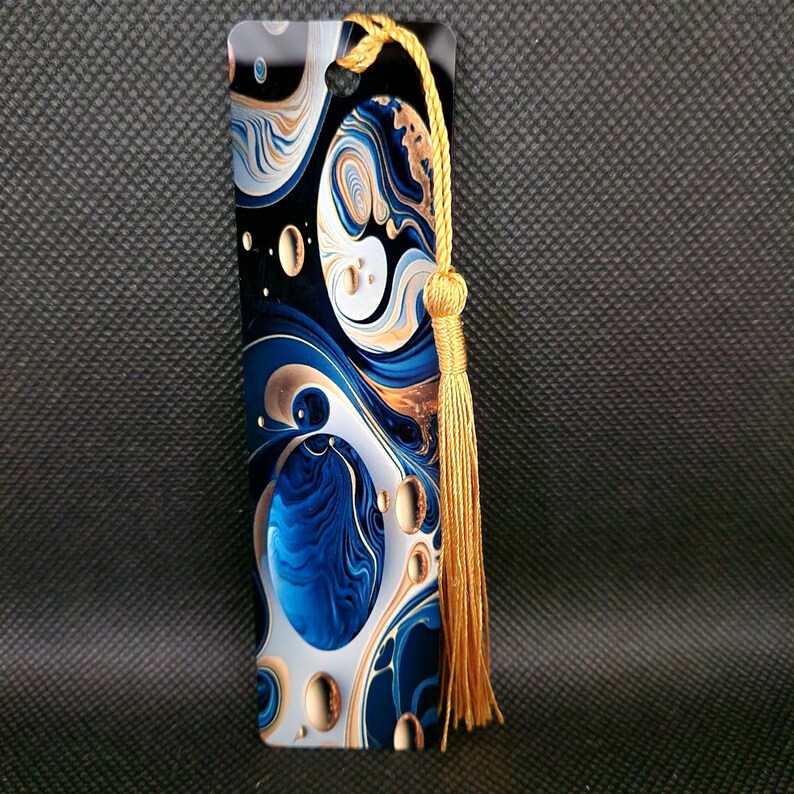 K&ouml;nnte beinhalten: Rechteckiger Lesezeichen mit einem wirbelnden abstrakten Design in Blau, Wei&szlig; und Gold. Das Lesezeichen hat eine goldene Quaste oben. Das Design zeigt eine Mischung aus organischen Formen und Mustern, die eine visuell ansprechende &Auml;sthetik erzeugen.