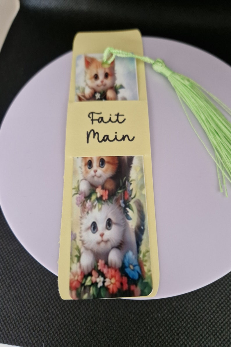 K&ouml;nnte beinhalten: Handgefertigter Lesezeichen mit K&auml;tzchen-Illustrationen. Der Lesezeichen hat einen hellgelben Hintergrund mit den Worten "Fait Main" in schwarzer Schrift. Eine gr&uuml;ne Quaste ist befestigt. Die Illustrationen zeigen K&auml;tzchen inmitten von Blumen.