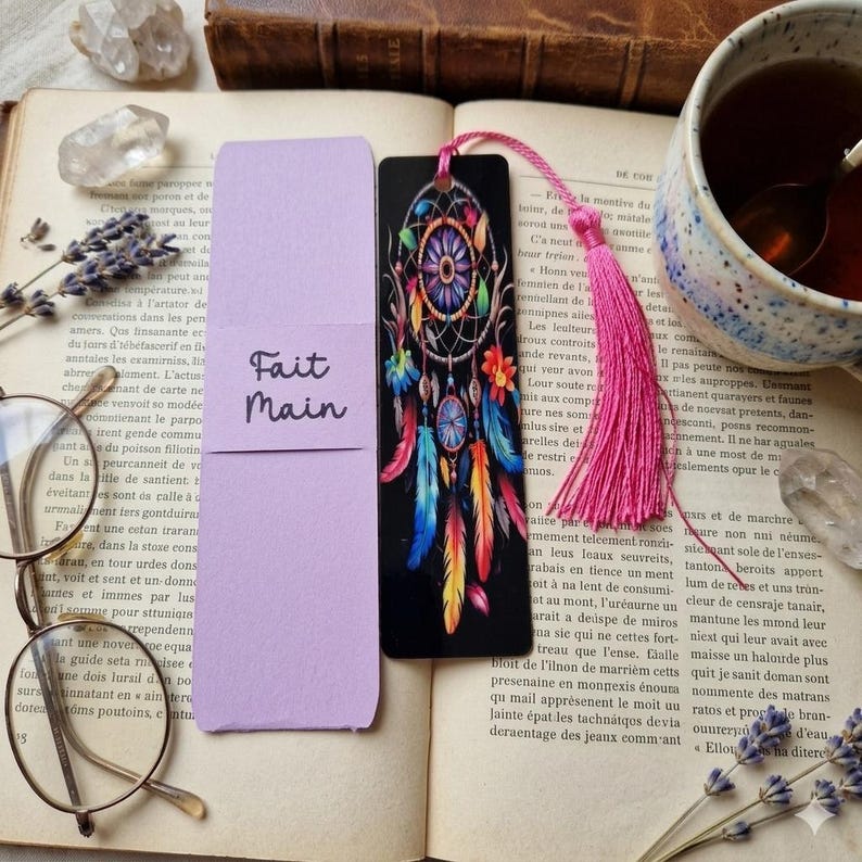 K&ouml;nnte beinhalten: Schwarzer Lesezeichen mit einem Traumf&auml;nger-Design und bunten Federn, neben einem lavendelfarbenen Lesezeichen mit dem Text "Fait Main". Eine rosa Quaste h&auml;ngt am Traumf&auml;nger-Lesezeichen.