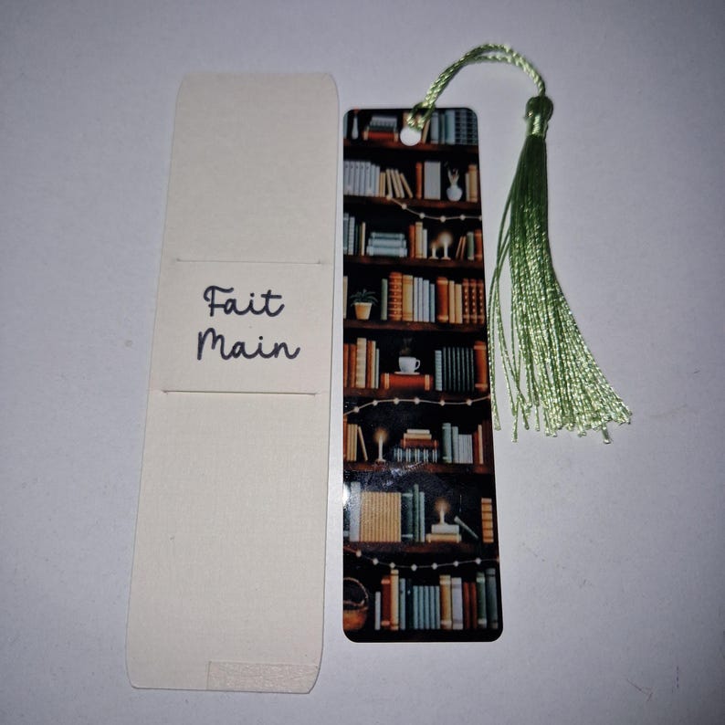 K&ouml;nnte beinhalten: Ein handgefertigtes Lesezeichen mit einem B&uuml;cherregal-Design und einer gr&uuml;nen Quaste. Das Lesezeichen zeigt eine detaillierte Illustration eines B&uuml;cherregals voller B&uuml;cher, Lichterketten und einer Kaffeetasse. Die Worte "Fait Main" sind auf der dazugeh&ouml;rigen Karte aufgedruckt.