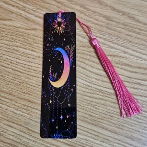 K&ouml;nnte beinhalten: Schwarzer Lesezeichen mit einem Himmelsdesign, das einen Mond in Blau, Gelb und Rosa zeigt. Das Lesezeichen hat eine rosa Quaste und ein Sonnen- und Sternenmuster. Das Lesezeichen ist rechteckig und hat eine gl&auml;nzende Oberfl&auml;che.