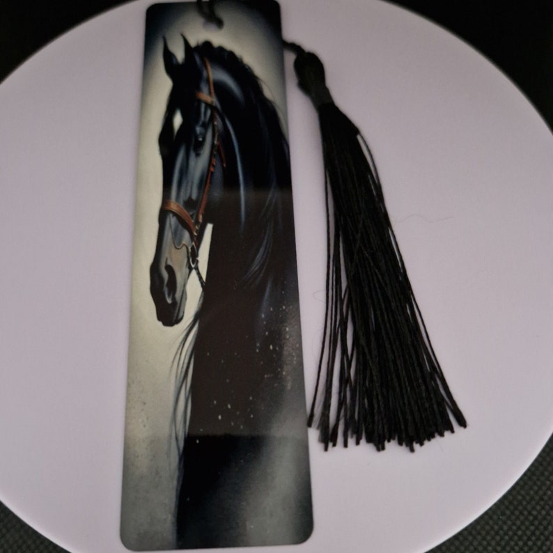 K&ouml;nnte beinhalten: Rechteckiger Lesezeichen mit einer Illustration eines schwarzen Pferdekopfes und einem braunen Zaumzeug. Der Hintergrund ist grau und wei&szlig;. Eine schwarze Quaste ist oben befestigt. F&uuml;r Buchliebhaber.