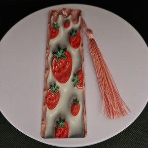 K&ouml;nnte beinhalten: Rechteckiger Lesezeichen mit Erdbeermuster. Das Lesezeichen hat einen wei&szlig;en Hintergrund mit roten Erdbeeren und gr&uuml;nen Bl&auml;ttern. Eine rosa Quaste h&auml;ngt oben. Das Lesezeichen ist etwa 15 cm lang.