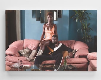 May include: A couple sitting on a pink sofa in a living room. The woman is wearing a peach satin dress and brown boots. The man is wearing a black and white tracksuit. There is a plant in the background.