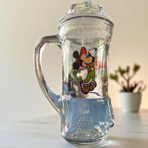 May include: Clear glass beer stein with a handle and a lid. The stein features a colourful image of Mickey Mouse in a red hat, green shirt, and brown shoes. The stein is sitting on a white surface.