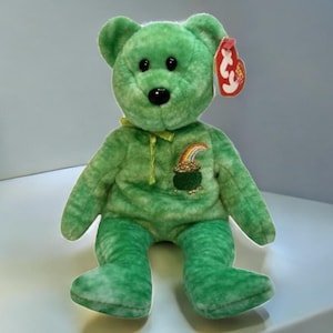 May include: Green plush beanie baby bear with a pot of gold and rainbow on its chest. The bear has black eyes and a black nose. The bear is sitting on a white surface. The bear has a red Ty tag attached to its ear.
