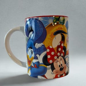Könnte beinhalten: Eine Keramik-Kaffeetasse mit weißem Henkel und rotem Rand, verziert mit Zeichentrickfiguren wie Donald Duck und Minnie Mouse. Das farbenfrohe Design ist detailliert und für Disney-Fans geeignet.