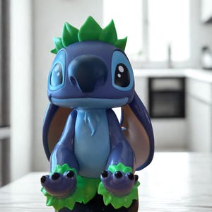 Puede incluir: Una figura azul y verde del personaje Stitch de la pel&iacute;cula de Disney Lilo & Stitch. La figura tiene orejas grandes, una nariz grande y detalles verdes en la cabeza y los pies. La figura est&aacute; sobre una base marr&oacute;n.