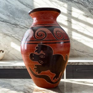 May include: A terracotta-colored vase with a dark brown rim and base. The vase features a black lizard design and decorative swirls. The vase is a decorative item for the home.
