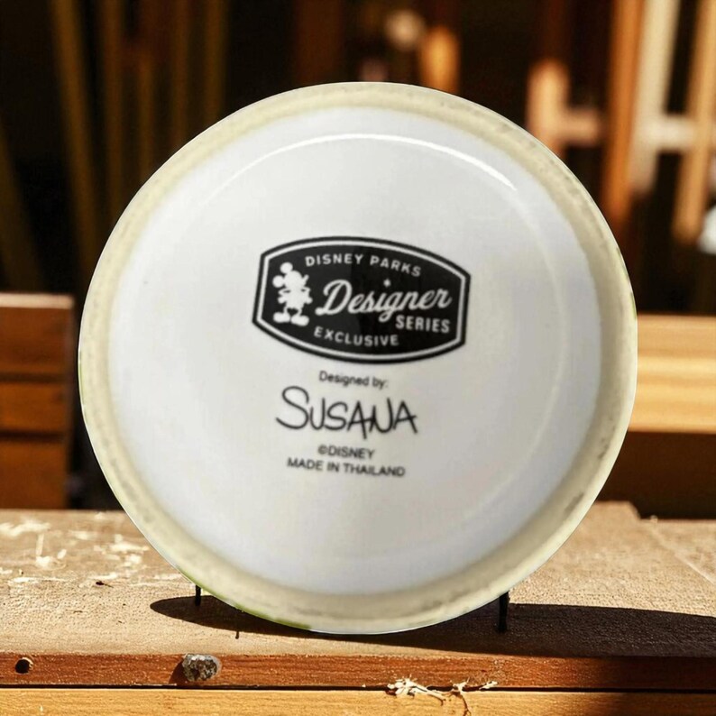 May include: A white ceramic mug base with a black and white Disney Parks Designer Series Exclusive logo. The mug is designed by Susana and made in Thailand. The mug sits on a wooden surface.
