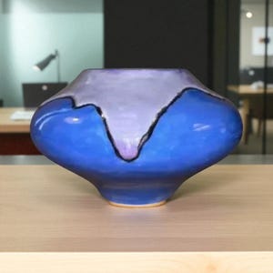 Puede incluir: Un jarrón de cerámica con una forma única y un acabado brillante. El jarrón presenta un degradado de colores, con una parte superior lila claro, un cuerpo azul profundo y un contorno negro. El jarrón está sobre una superficie de madera.