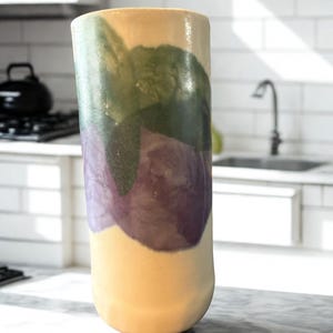 May include: A tall, cylindrical ceramic vase with a rounded base. The vase features a color palette of yellow, green, and purple, with the colors blending in an abstract pattern. The vase is displayed on a marble countertop in a kitchen setting.