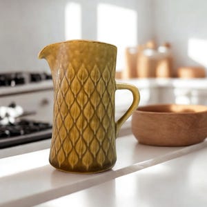 Puede incluir: Jarra alta de cerámica verde oliva con un patrón de hojas texturizadas. La jarra tiene un asa y un pico para verter. Un pequeño cuenco de madera está cerca. La jarra está sobre una encimera blanca en un entorno de cocina.