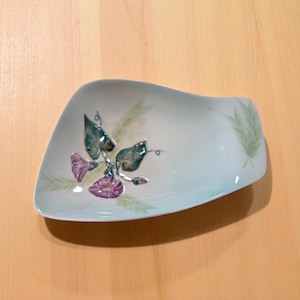 May include: A light blue, uniquely shaped ceramic dish. The dish is decorated with a floral design featuring purple flowers, green leaves, and delicate green stems. The dish is likely used for serving or display.