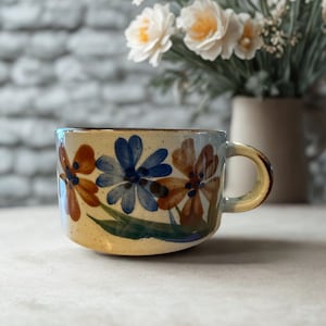 May include: A ceramic mug with a floral design. The mug features brown, blue, and orange flowers with green leaves against a cream-coloured background. The handle and rim are brown.