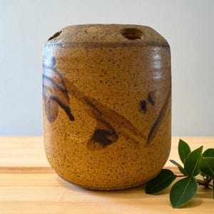 Peut inclure: Un vase cylindrique en céramique fait à la main, dans les tons bruns et beiges. Le vase présente une texture tachetée et des motifs abstraits brun foncé. Le dessus comporte trois ouvertures circulaires. Une branche de feuilles vertes repose sur la surface en bois.