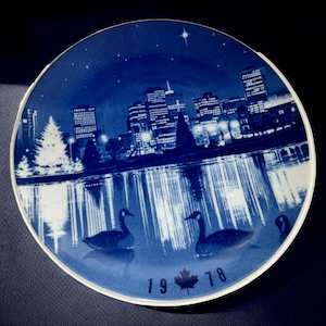 May include: A blue and white decorative plate depicting a city skyline at night, with a lake and geese. The plate includes the year 1978 and a white rim. The design is reminiscent of a winter scene.