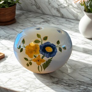 May include: A round, off-white ceramic flower frog with hand-painted yellow and blue floral designs. The frog has several holes on top for arranging flowers. The background is a marble countertop.