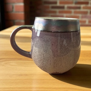 May include: A ceramic mug with a rounded body and a dark purple handle. The mug has a gradient colour scheme, transitioning from a light speckled purple at the base to a darker purple with iridescent accents near the rim. The interior is white.
