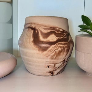 May include: A beige ceramic vase with a marbled brown pattern. The vase has a wide base, a slightly curved body, and a narrower top. The design resembles swirling earth tones. The vase is a decorative item.