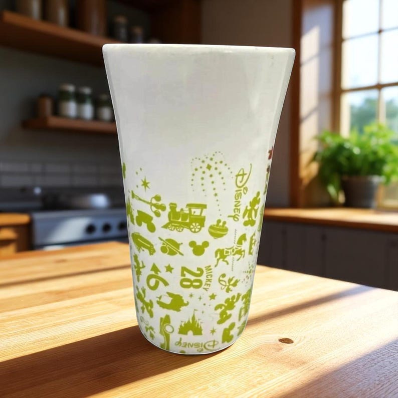 May include: A white ceramic tumbler with a green Disney-themed pattern. The design includes a train, Mickey Mouse, and other iconic images. The tumbler is on a wooden surface.