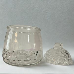 May include: A clear glass honey jar with the word HONEY embossed in capital letters around the side. The jar has a matching clear glass lid featuring a textured, raised honeycomb pattern and a small knob handle on top for easy removal.