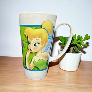 Puede incluir: Taza alta de cerámica blanca con una imagen de dibujos animados de Campanilla con cabello rubio y un vestido verde. La taza tiene un asa curva y un borde verde y azul. Una pequeña planta suculenta en maceta está al lado de la taza.