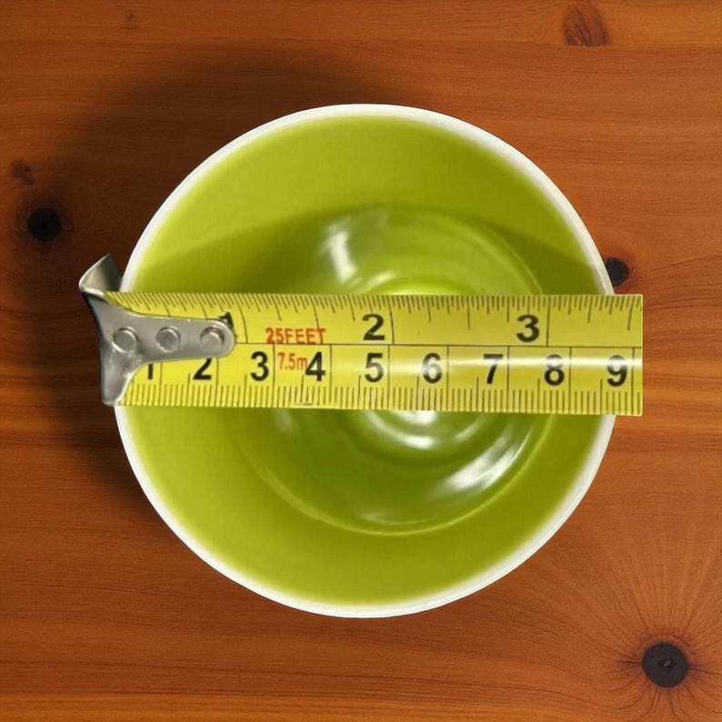 May include: A lime green ceramic bowl with a white rim is measured with a yellow tape measure. The tape measure shows the bowl's diameter is approximately 7 inches. The bowl sits on a wooden surface.