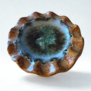 Puede incluir: Un cuenco de cerámica hecho a mano con una forma única y orgánica. El cuenco presenta un borde ondulado y una paleta de colores marrón, azul y verde. El interior tiene un aspecto texturizado y vidrioso, y el exterior tiene un acabado moteado y terroso.