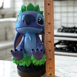 Puede incluir: Una figura azul y morada de un personaje de dibujos animados con orejas grandes y detalles verdes. La figura mide aproximadamente 20 cm de altura y se apoya sobre una base negra. El personaje tiene ojos grandes y una expresi&oacute;n amigable.