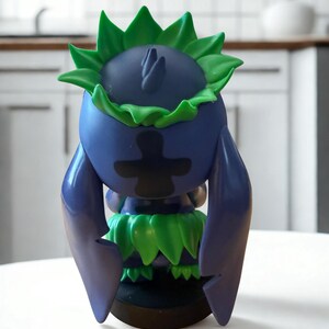 Puede incluir: Figura azul y verde con un dise&ntilde;o &uacute;nico. La figura tiene una cabeza redonda con detalles verdes en forma de hojas y un dise&ntilde;o negro en forma de cruz en la parte delantera. Est&aacute; sobre una superficie blanca.