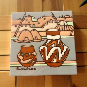 May include: Square ceramic tile featuring a Southwestern design. The tile depicts a desert landscape with adobe structures, ladders, and two pottery vessels in shades of brown, orange, and white against a gray background. The tile is signed "Taissedre 06".