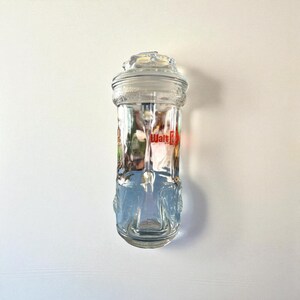 May include: A clear glass jar with a lid, featuring the text "Walt" in red. The jar has a decorative base and a detailed lid. The jar is cylindrical and appears to be empty.