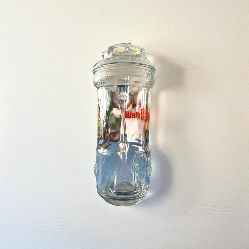 May include: A clear glass jar with a lid, featuring the text "Walt" in red. The jar has a decorative base and a detailed lid. The jar is cylindrical and appears to be empty.