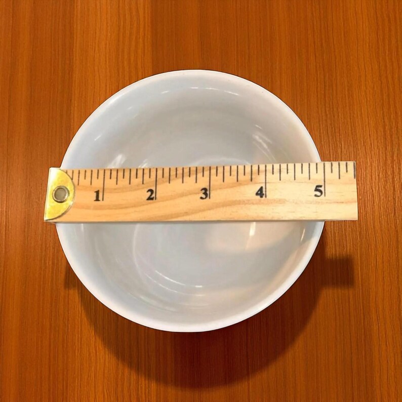 May include: A white ceramic bowl with a wooden ruler placed across the top. The ruler shows measurements in inches, with the bowl appearing to be approximately 5 inches in diameter. The bowl is round and has a smooth, glossy finish.