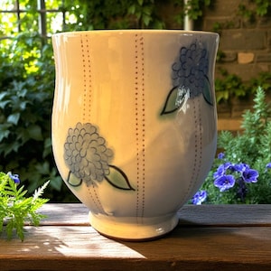 May include: Cream ceramic mug with blue floral designs and green leaves. The mug has vertical dotted lines and a flared top. The mug is on a wooden surface, with greenery in the background.